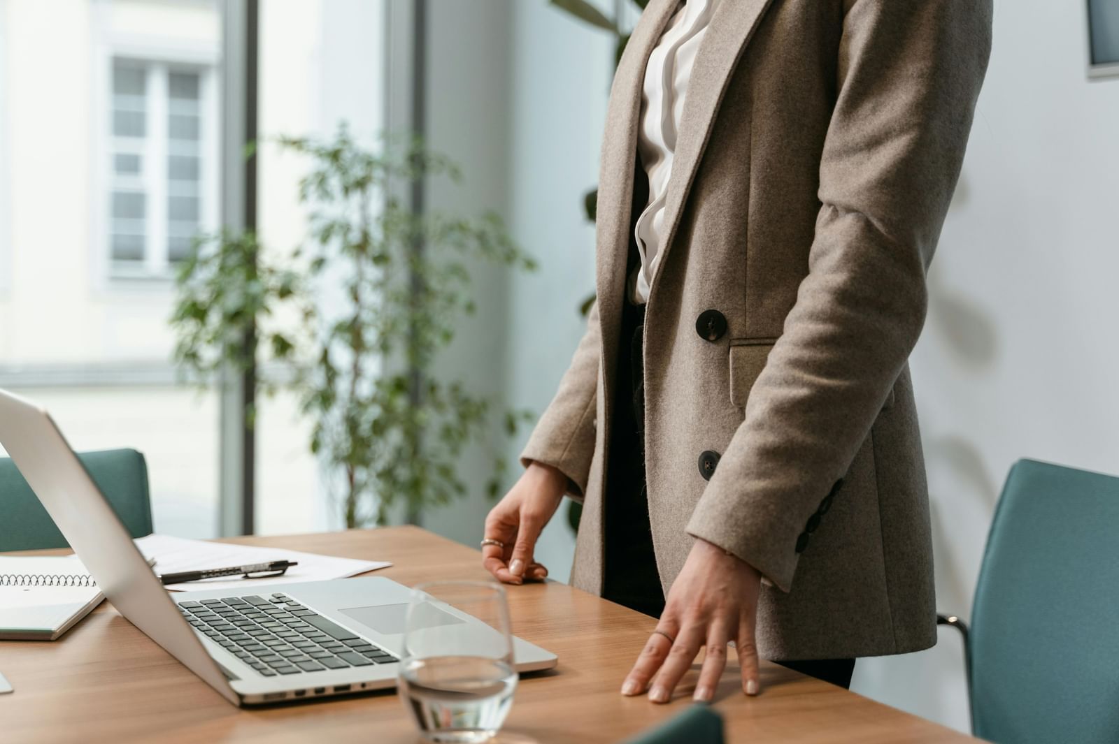 Person in a blazer standing at a desk with a laptop, notebook, and glass of water, in a bright, modern office setting