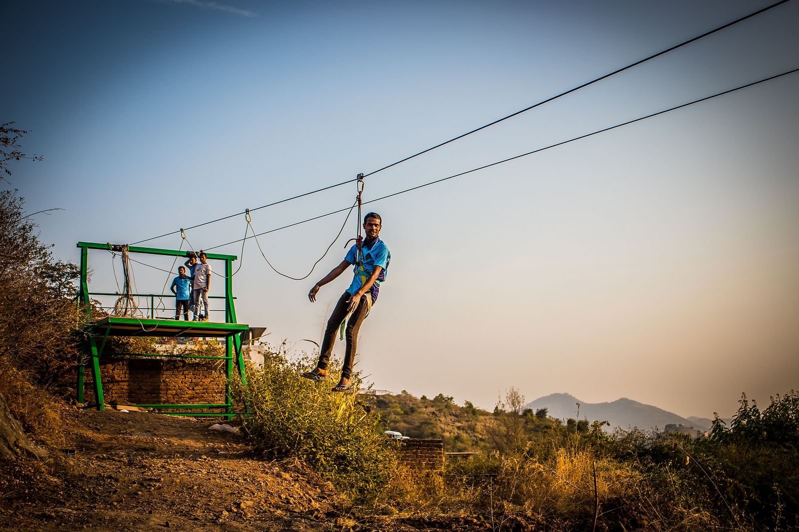 A person zip-lining across a scenic landscape with hills in the background.- Bamboo Saa Resort & Spa, Udaipur