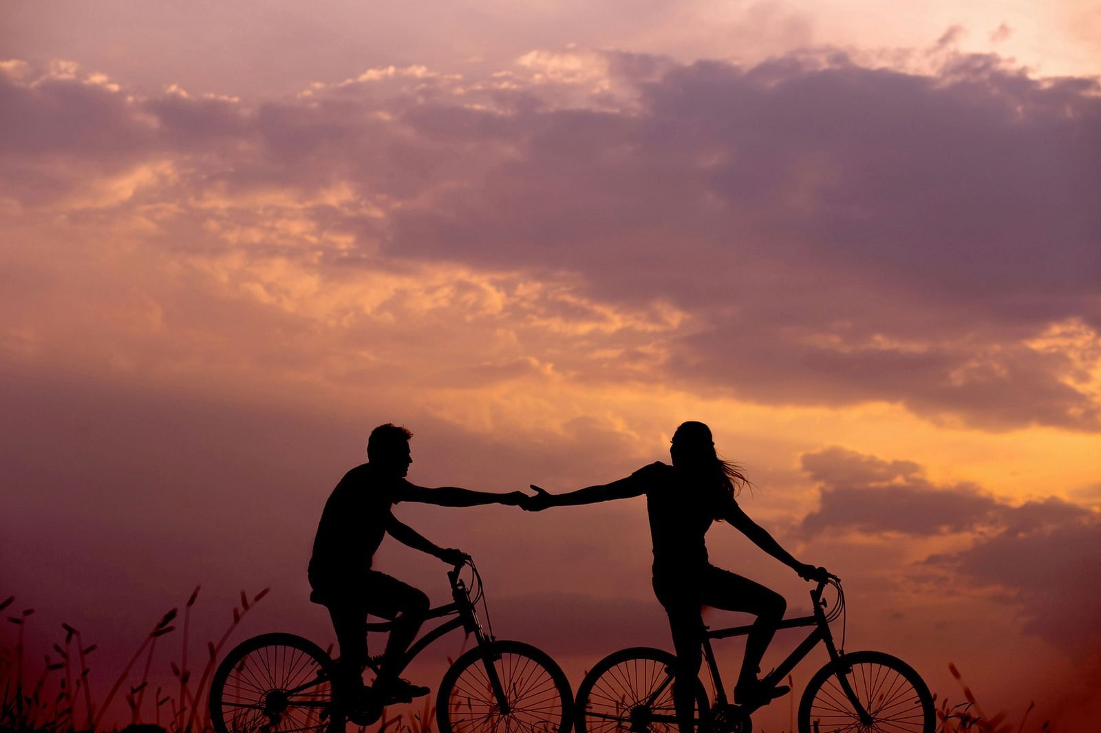 Silhouettes of two people on bicycles, holding hands, against a dramatic sunset sky with clouds. 