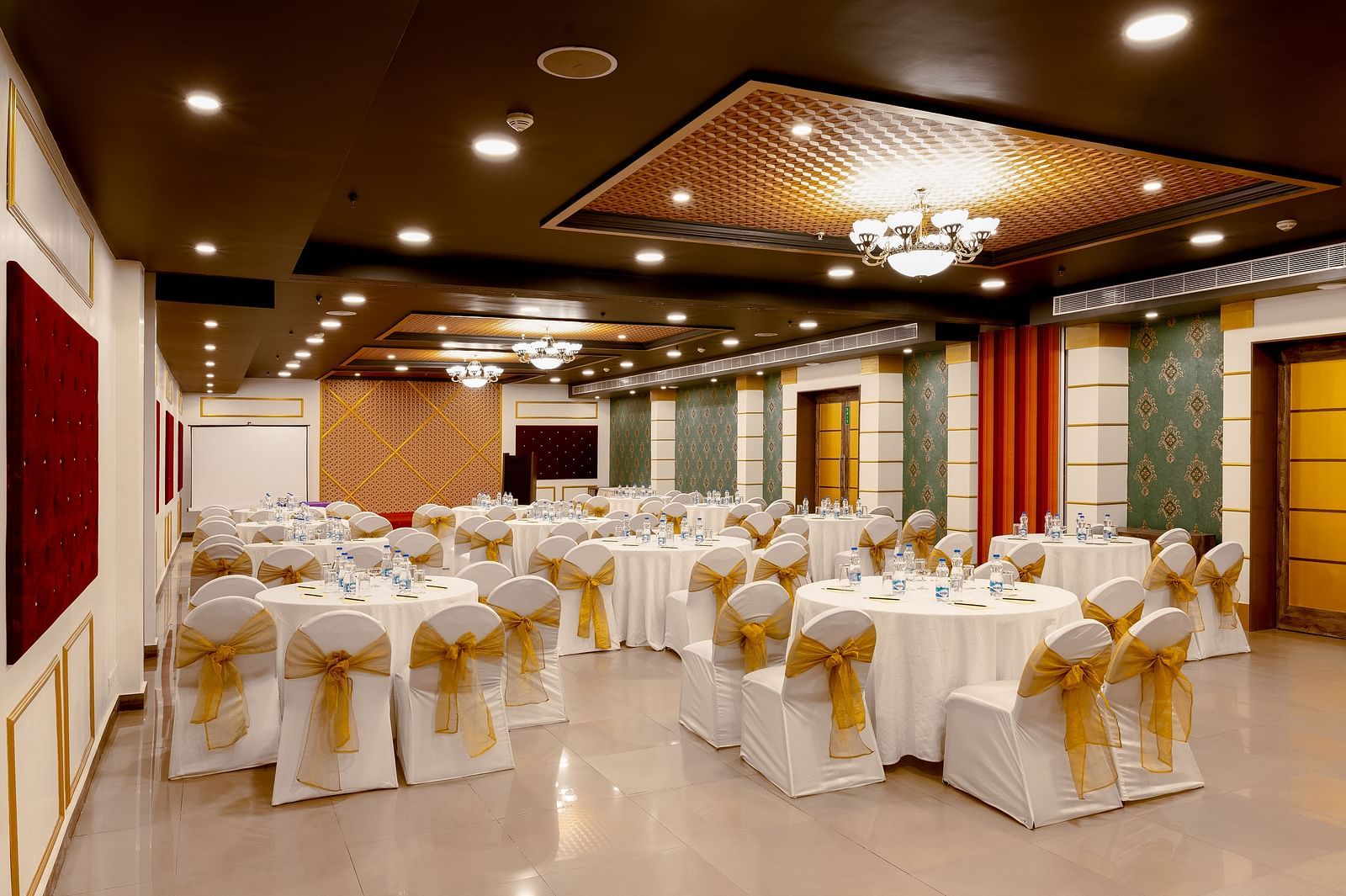 A spacious banquet hall with round tables covered in white cloths and gold chair covers. The hall has a modern design with dark wood accents and a crystal chandelier.