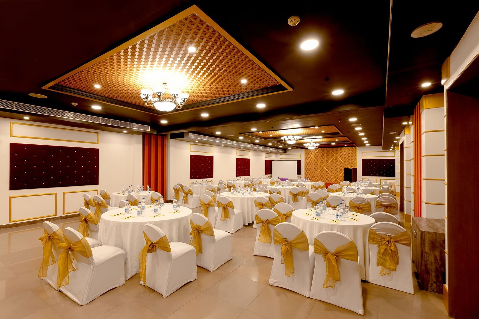 A spacious banquet hall with round tables covered in white cloths and gold chair covers. The hall has a modern design with dark wood accents and a crystal chandelier.