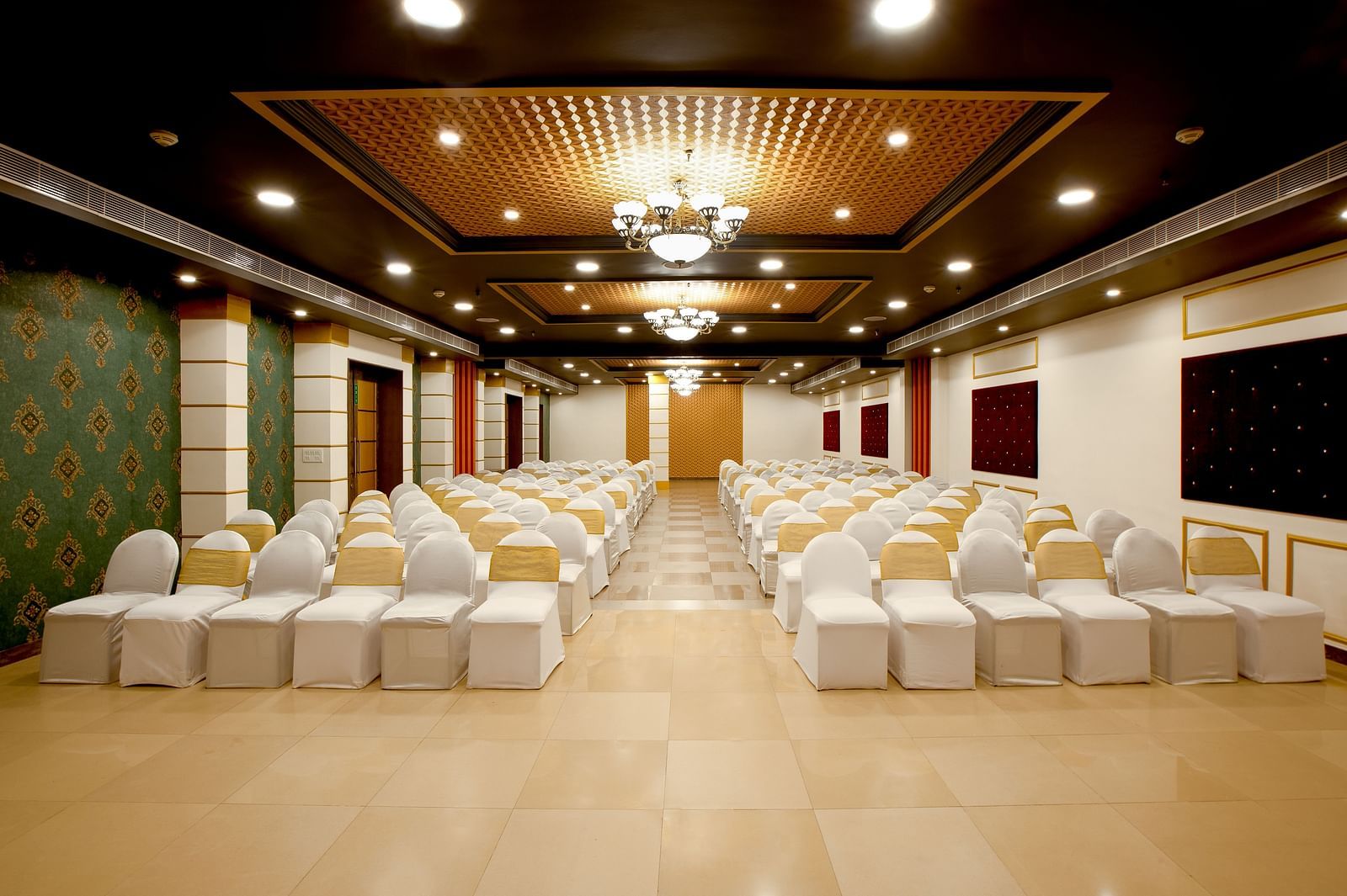 A spacious banquet hall with chairs covered in white cloths and gold chair covers. The hall has a modern design with dark wood accents and a crystal chandelier.