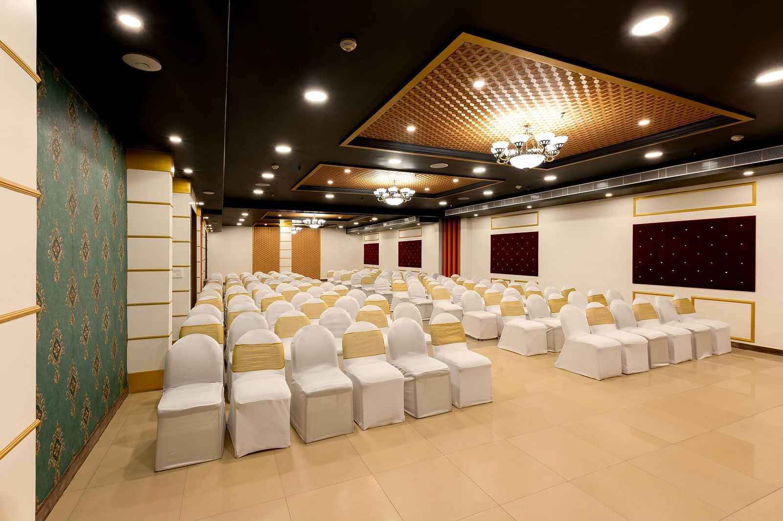 A spacious banquet hall with  chairs covered in white cloths and gold chair covers. The hall has a modern design with dark wood accents and a crystal chandelier.