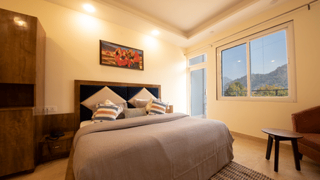 A hotel room, featuring a large bed with a grey duvet and a wooden frame, a painting on the wall, and a window with a view of hills | Perfectstayz Value Hills