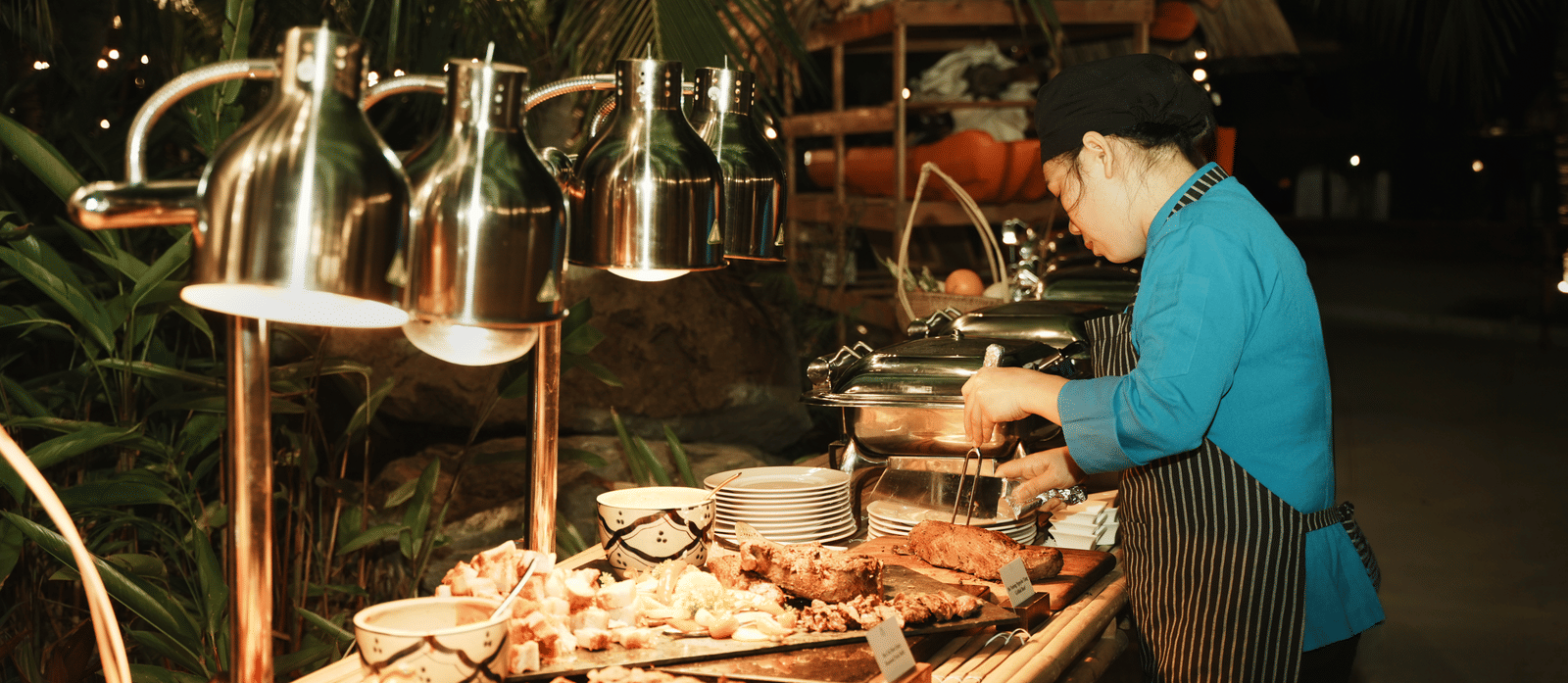 A woman at the barbecue table checking whether the meat is cooked. The table is filled with cooked meat, vegetables, and cooking utensils at Albu Resorts, Vietnam.
