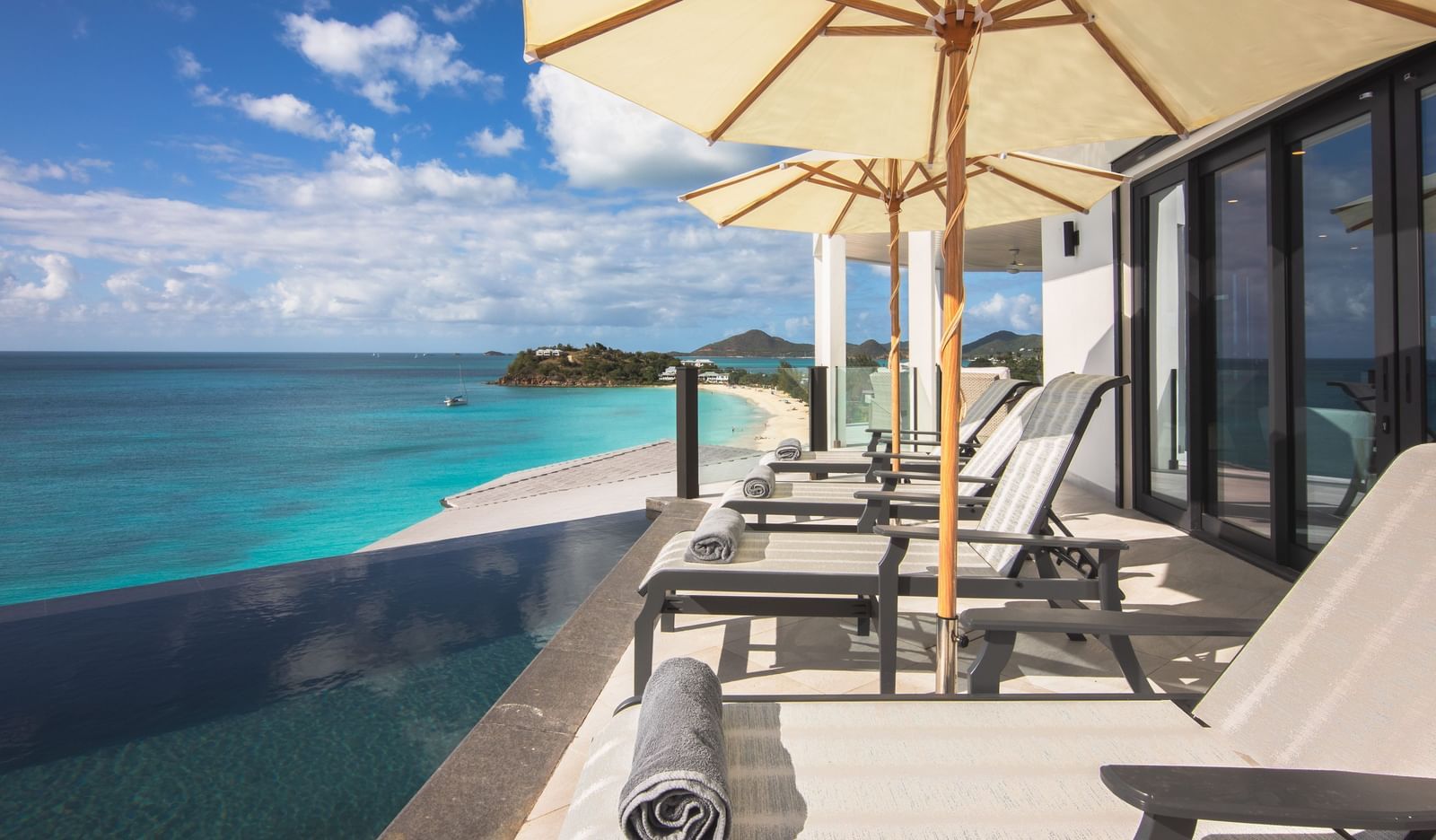 A patio with lounge chairs and umbrellas overlooks a white sand beach and turquoise ocean under a blue sky.