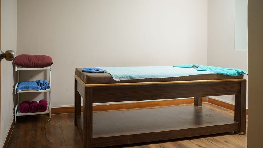 A wooden massage table in a room with light-coloured wooden flooring and white walls, with some folded towels on a shelf at Orsino Spa Resort.