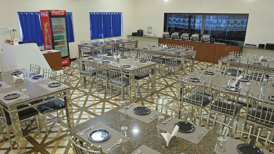 A wide shot of the restaurant at VITS Select Diamond Resort, Sasan Gir. The room is filled with tables and steel chairs, a buffet counter, and a Coca-Cola refrigerator.