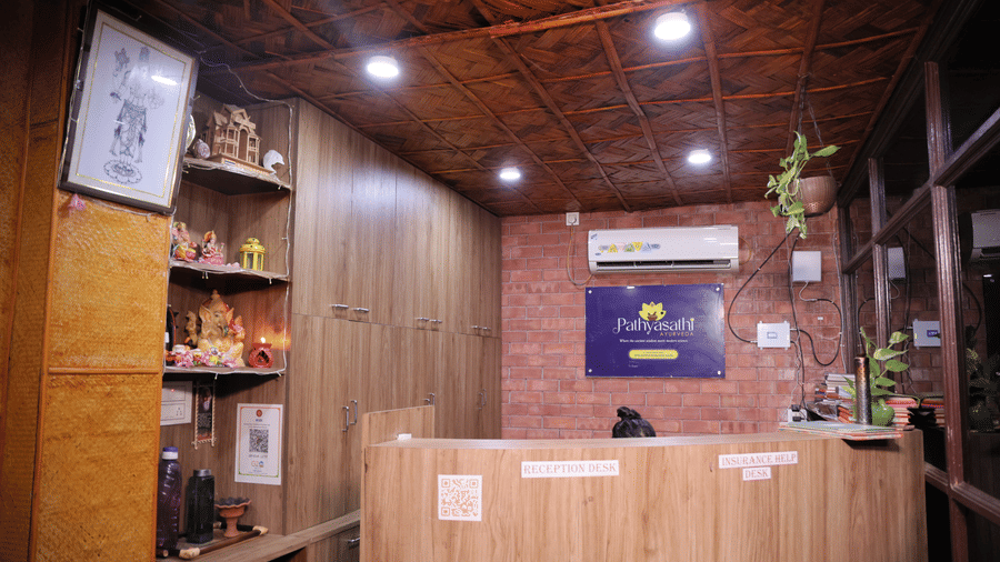 A wooden reception desk under a panelled ceiling with inset lights  and shelves with items, at Shophori Wellness Retreat, Bolpur.