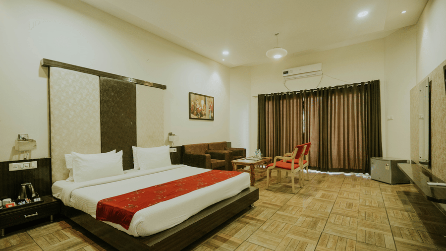 A bedroom at TGI Jahaj Mahal, Mandu, with a king size bed with a red runner, side tasbles, and a separate seating area with chairs and sofa.