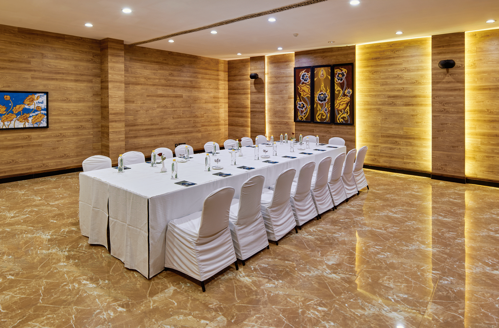 A modern conference room set up in a hollow square configuration with a long table covered in a white cloth and white slipcovered chairs, featuring marble floors and wood-panelled walls with back-lit accents.