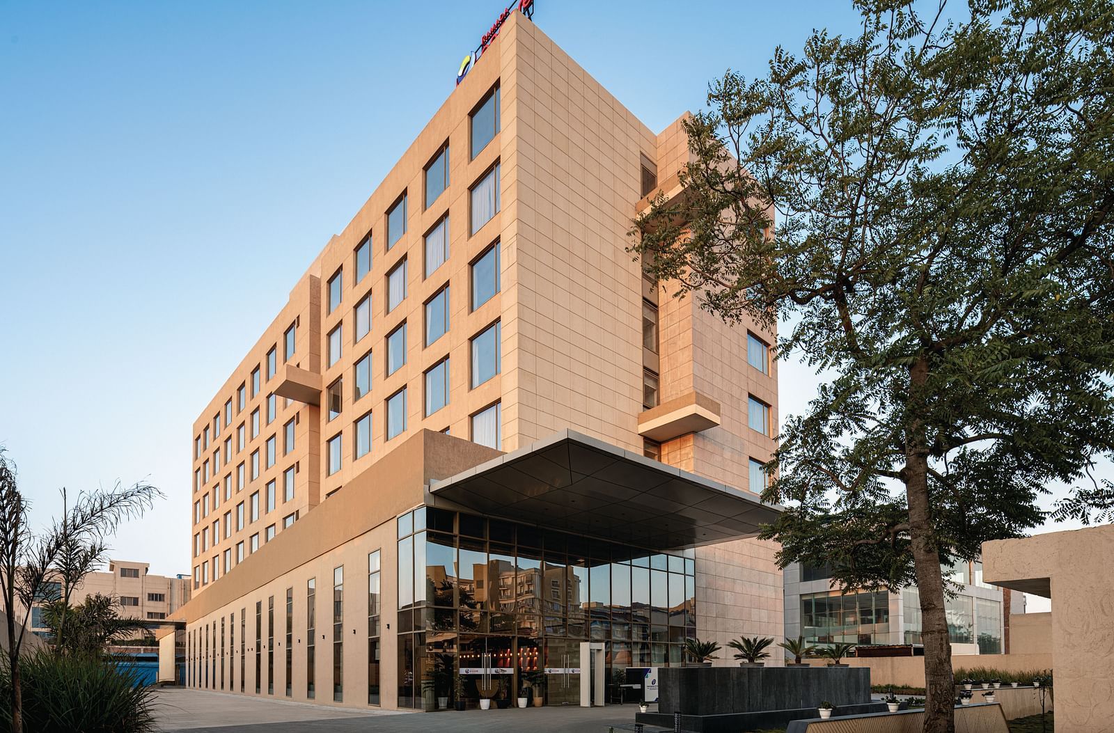Ramada Encore by Wyndham, Bhiwadi | Exterior daytime view of a tall, modern hotel building with a geometric facade. A black awning provides shade over an outdoor patio with seating. Trees are visible in the foreground.