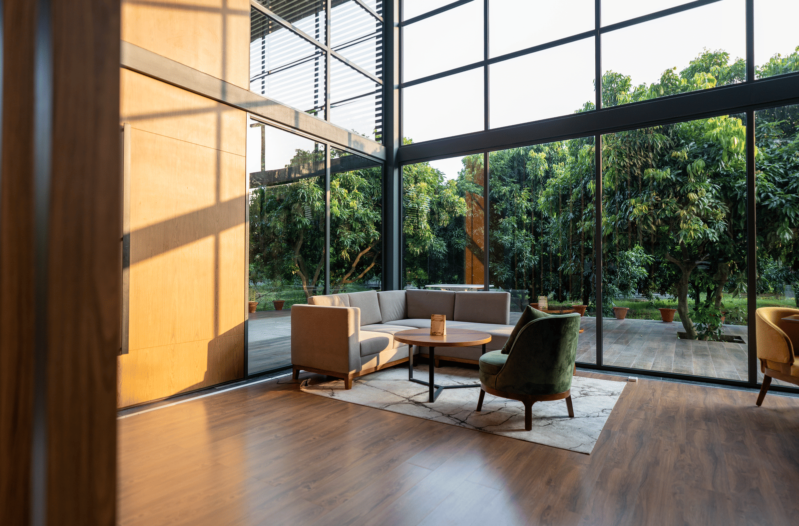 A sun-lit seating area at Wyndham Garden featuring a sofa, an armchair, and a low coffee table on a rug, framed by floor-to-ceiling windows with views of dense greenery.