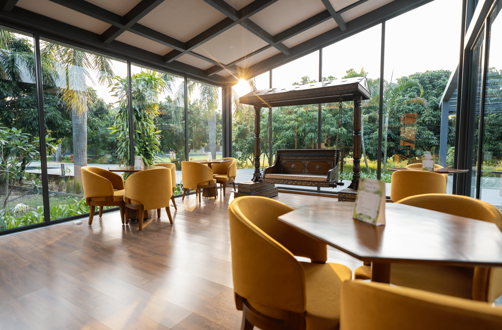A bright glass-walled lobby at Wyndham Garden with a wooden floor, round tables with upholstered armchairs, a central wooden swing bench, and views of lush greenery outside.