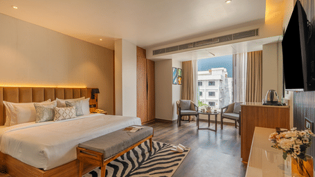 A bedroom featuring a king size bed, wall-mounted TV, bench seat, zebra-print rug, and city views at Perfectstayz Premium Mahadev Regency, Katra.