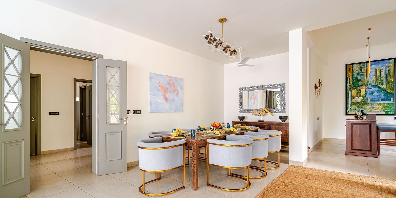 Elegant indoor dining area with a wooden table set for a meal, surrounded by plush gray chairs with gold accents, decorative artwork, a mirror, and modern lighting in a spacious, well-lit room - Lindsay Manor