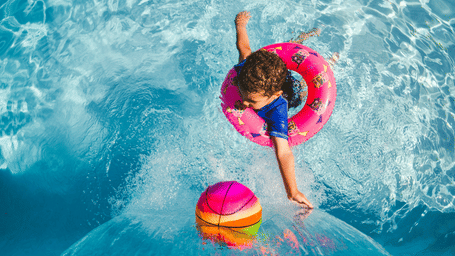 Young child floating in a swimming pool with a colourful inflatable ring and ball, enjoying a playful moment in the water.