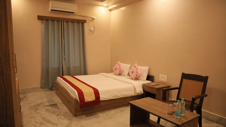 A bedroom featuring a king size bed with a red-striped runner, a small table, a wooden chair, air-conditioner, and window curtains at Shophori Wellness Retreat, Bolpur.