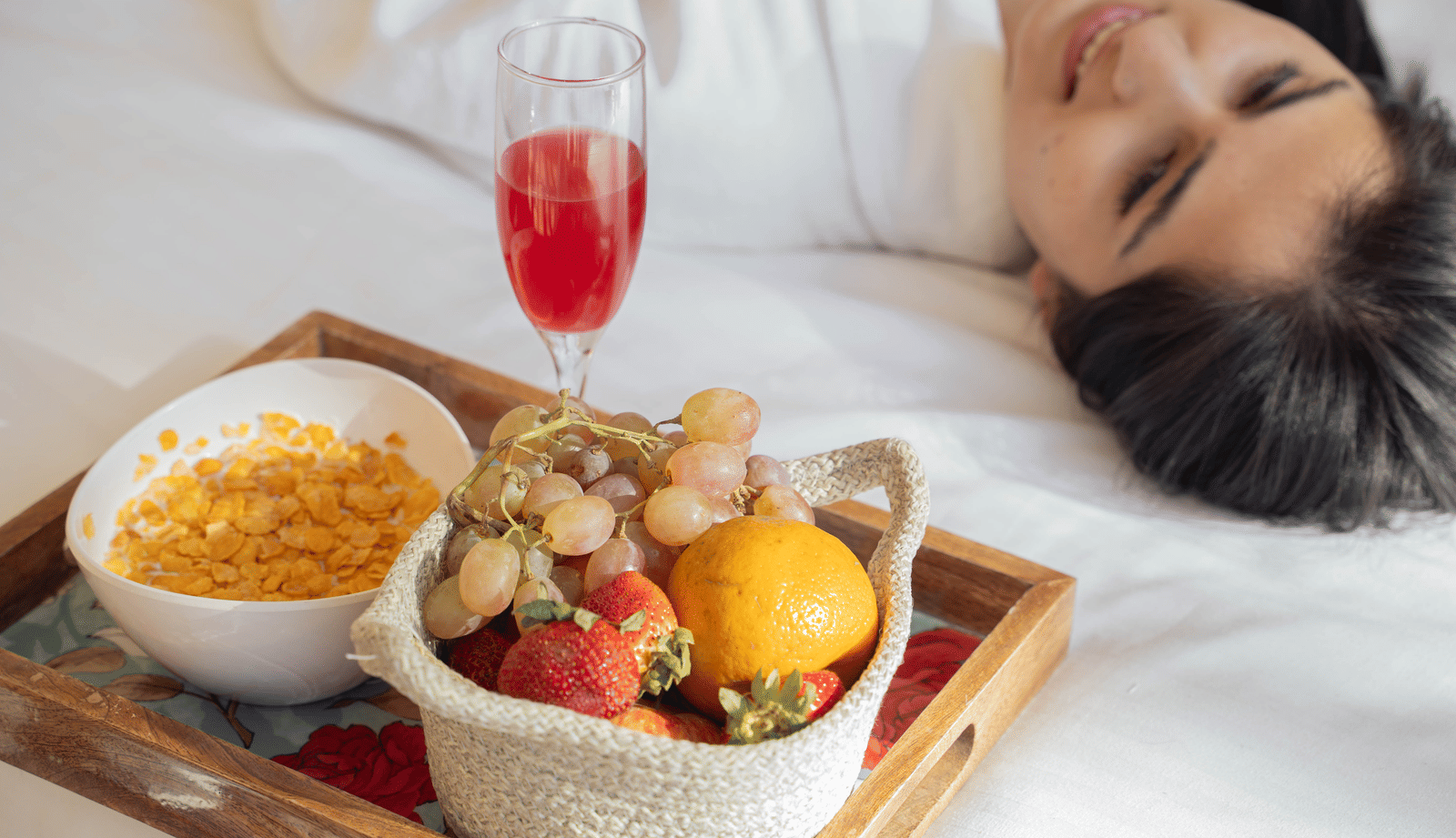 A tray on a bed with a bowl of cornflakes, a basket of fruits and a glass of red beverage, with a person lying beside it at Perfectstayz Value Shimla (Namah Retreat).