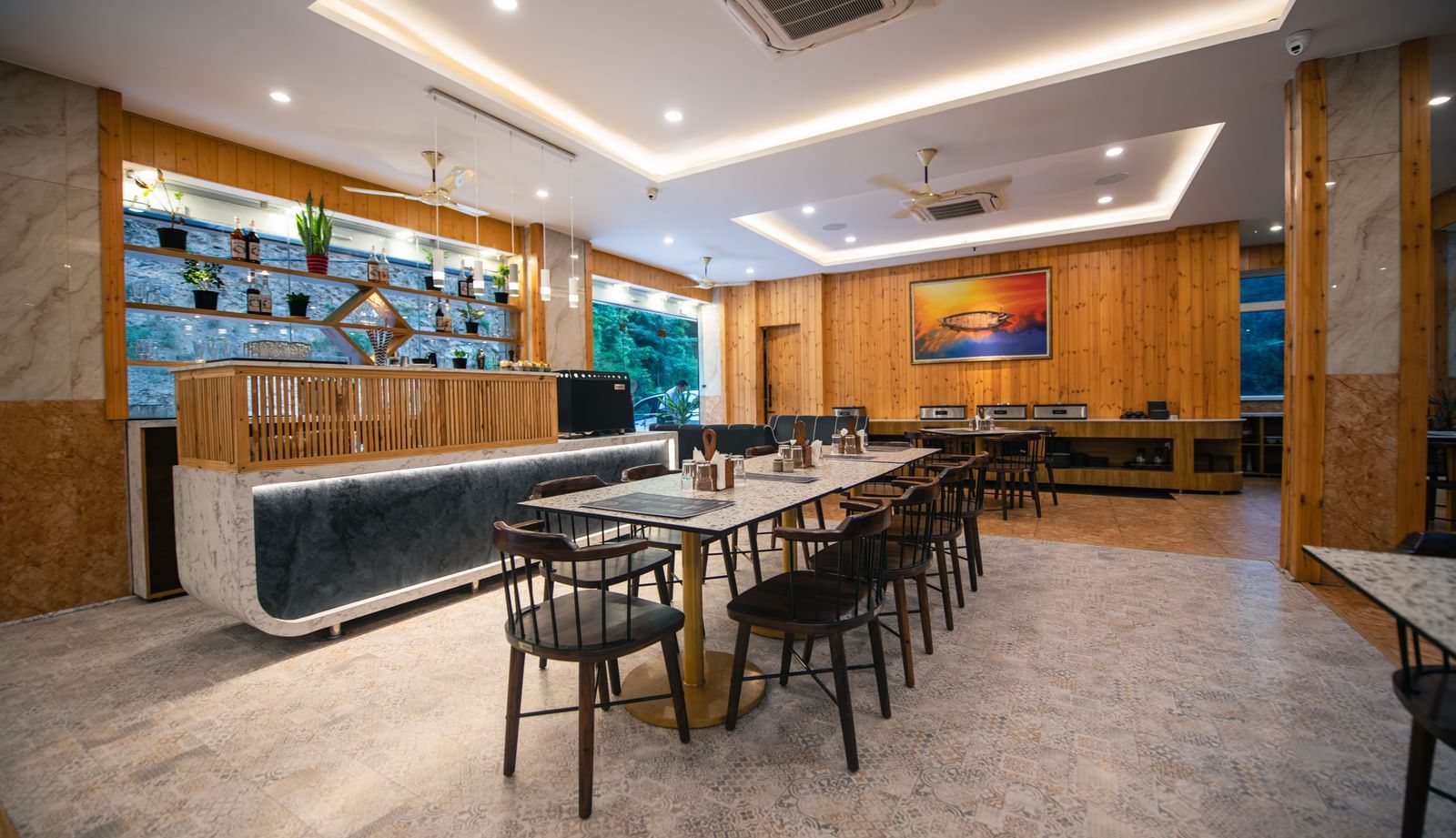 A restaurant at The Tattva Devaprayag with a long wooden table with black chairs and a bar.