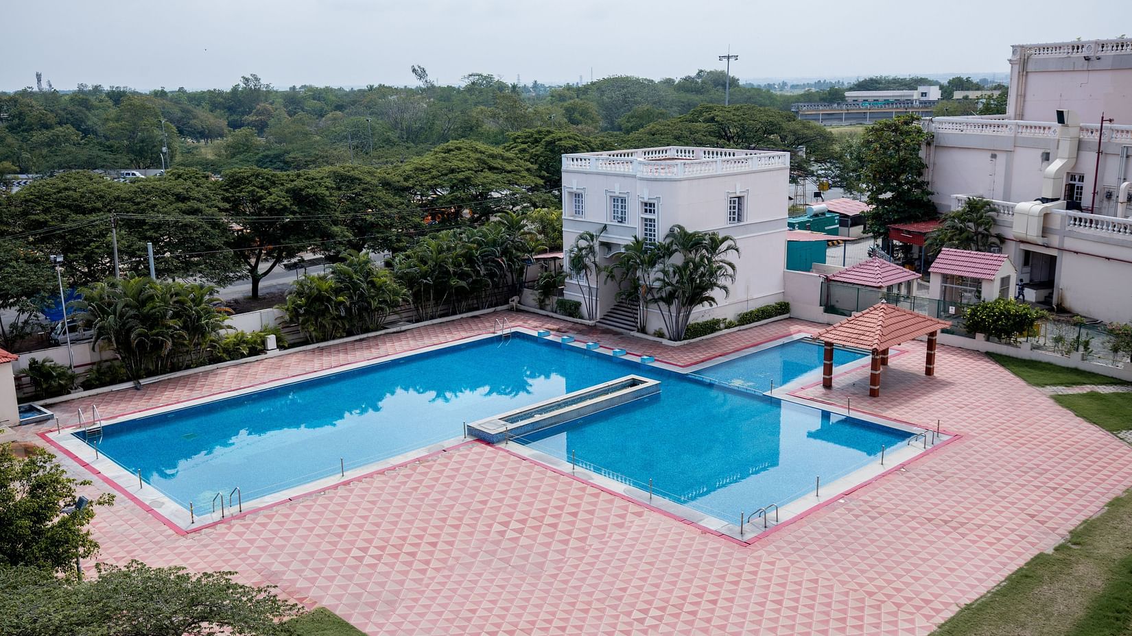 A large outdoor swimming pool with clean water, surrounded by a tiled deck, nearby buildings, and trees under a cloudy sky at Jenneys Residency