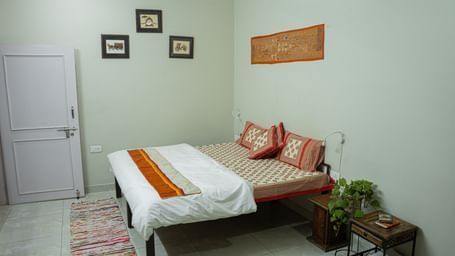 A simple bedroom with a double bed, white bedding, orange throw, wall frames and rug at Jaypore360 Homestay in Jaipur.