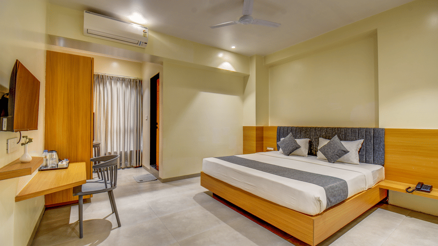 A well-lit Deluxe Room featuring a plush bed facing the TV on the wall below which is a work desk near the wardrobe