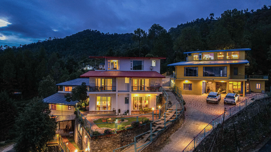 An aerial view of Adrushya Estates, a hill resort in Mukteshwar, on a hillside with lights on and trees around during evening time.