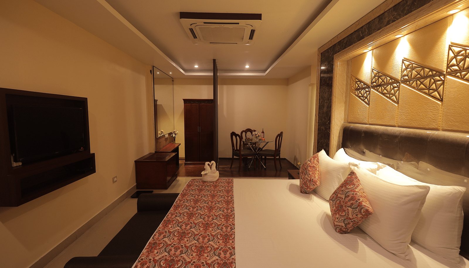 An image of a bedroom with bedside table, a set of table and chairs, flat TV screen, and AC- Sathyam Grand Resort