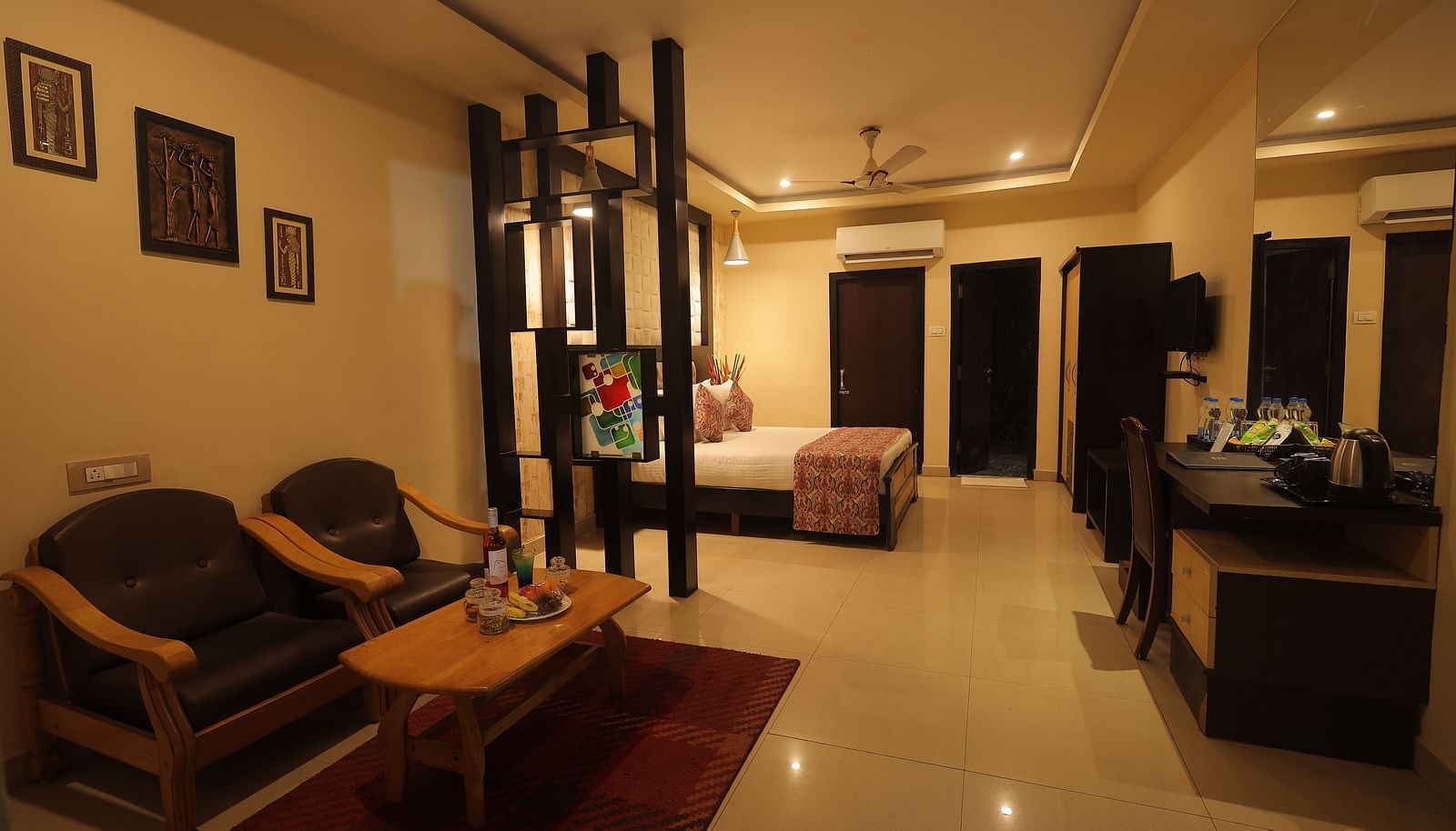 An image of a bedroom with bedside table, a set of table and chairs, two sofa seats, flat TV screen, and AC - Sathyam Grand Resort