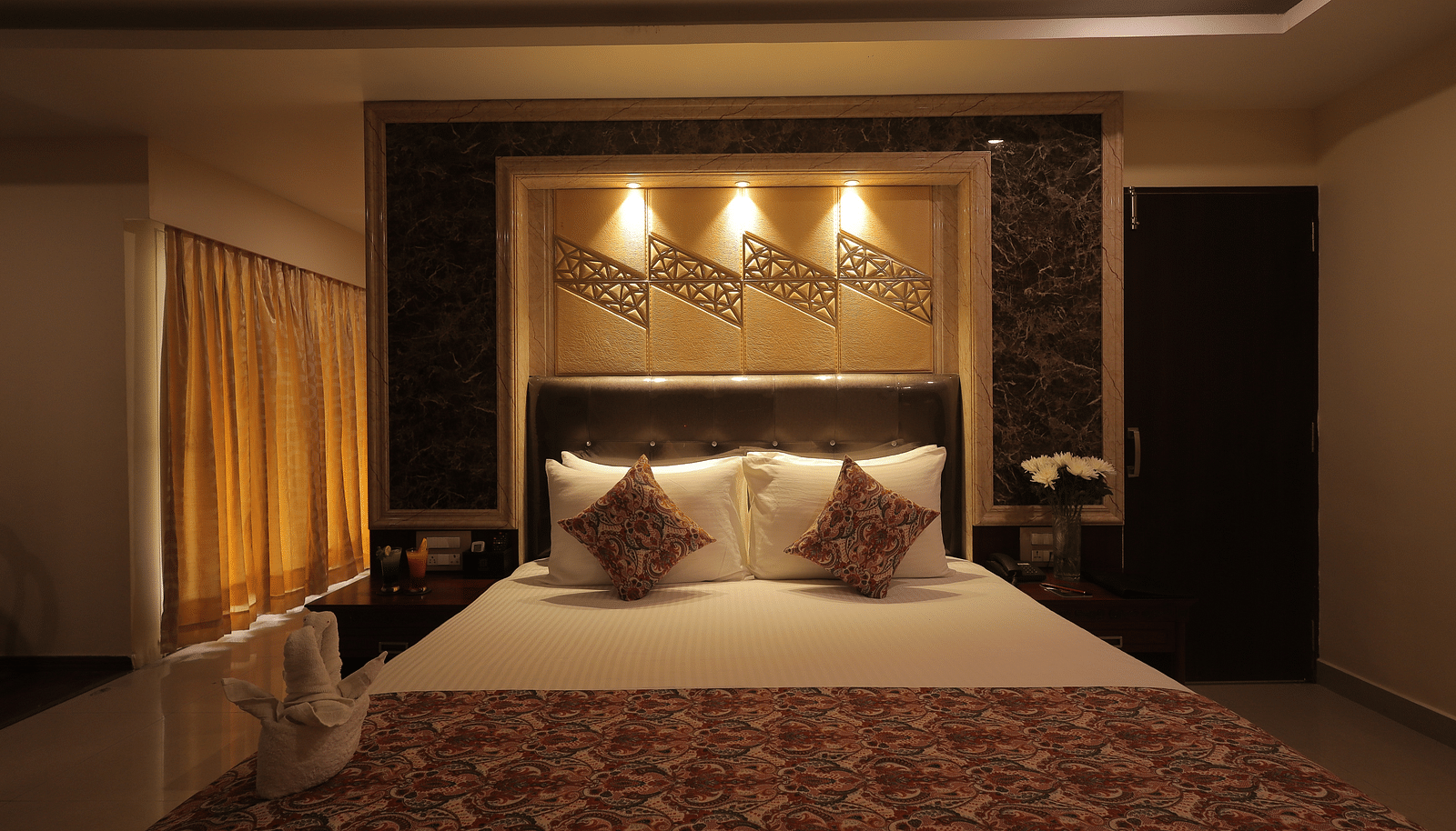 An image of a bedroom with bedside table, a set of table and chairs, flat TV screen, and AC - Sathyam Grand Resort