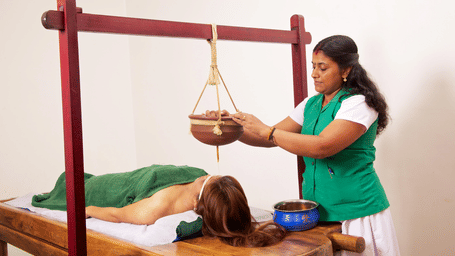 A therapist adjusting a hanging wooden pot over a person's forehead during a traditional Ayurvedic oil treatment | Le Pondy