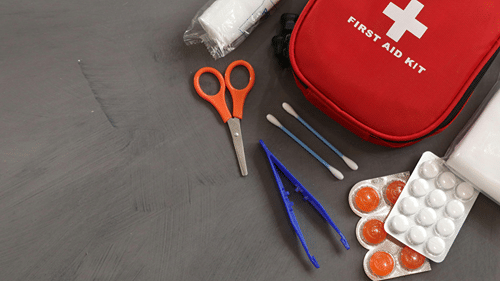 A red first aid kit pouch resting on a flat surface alongside medical supplies including scissors, tweezers, swabs, and pill blister packs.