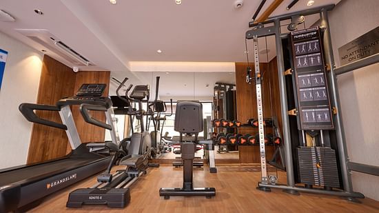 A well-equipped hotel gym features a treadmill, elliptical machine, weight bench, and a cable machine with various weight plates, all on a wooden floor. - Iris Sarovar Portico, Sector 85, Gurugram