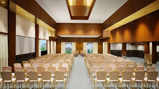 A well-lit event hall with chairs arranged neatly facing a central stage, featuring modern wooden interiors, windows offering natural light, and a sleek, polished floor - Uday Backwater Resort