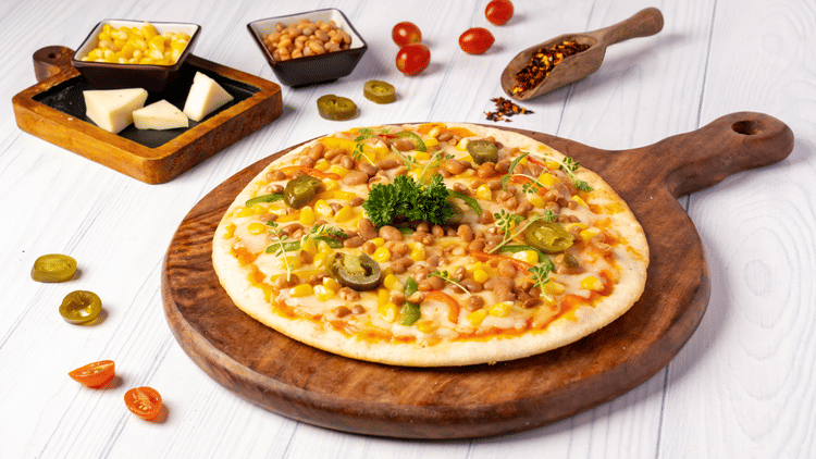 A round pizza on a wooden board is surrounded by a small bowl of corn, cheese wedges, and cherry tomatoes.