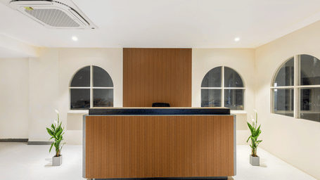 A wooden reception desk, in a white lobby area flanked by potted plants and arched window frames at Monday Hotels & Suites Madhapur Hyderabad.