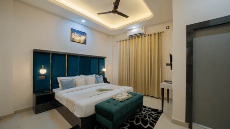 A bedroom with a king size bed, bedside tables, curtains, ceiling fan, wall art, and a bench at Perfectstayz Value Aalaya Suites, Haridwar.