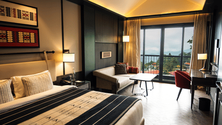 Hotel room featuring double bed with striped runner, sofa, desk with red chair, and large glass doors leading to balcony.