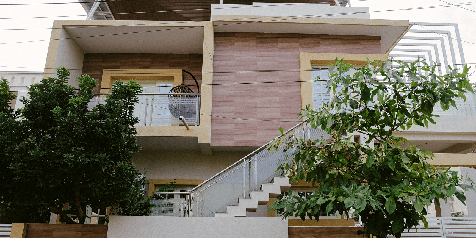 A modern two-story house with a combination of light-colored and wood-paneled exterior, featuring a staircase leading to an upper balcony.