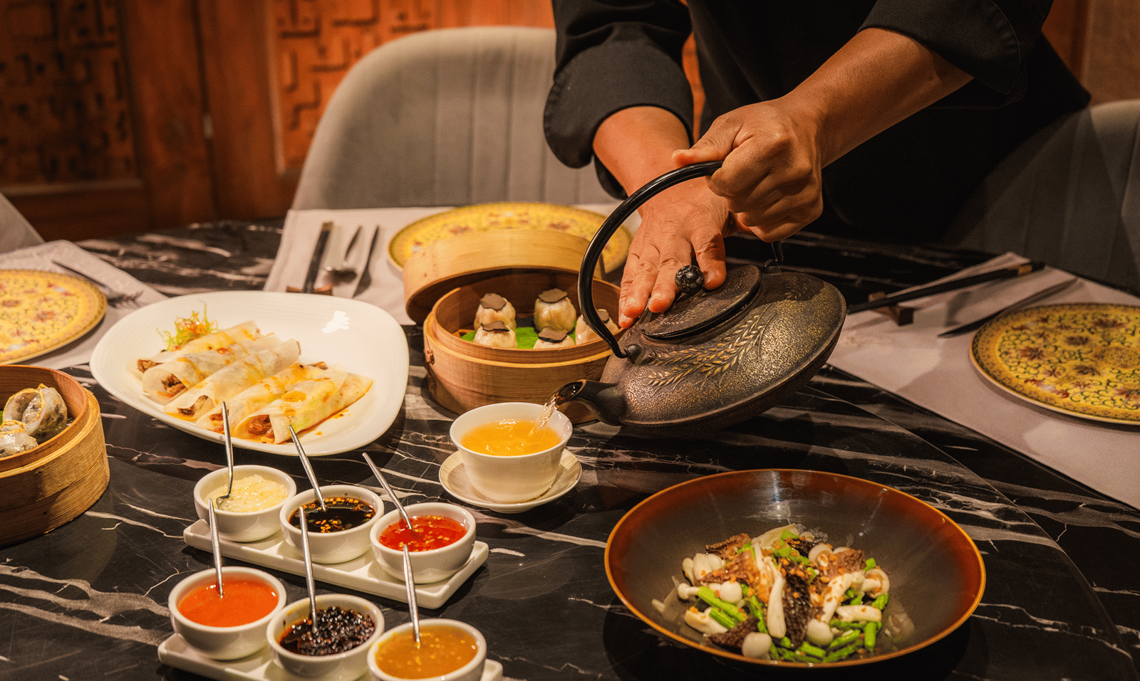 A chef pouring Chinese tea from a cast-iron teapot beside truffle dim sum, a morel stir-fry and dipping sauces at Jade, The Claridges New Delhi