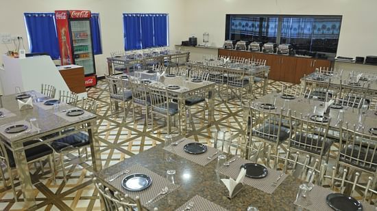 A wide shot of the restaurant at VITS Select Diamond Resort, Sasan Gir. The room is filled with tables and steel chairs, a buffet counter, and a Coca-Cola refrigerator.