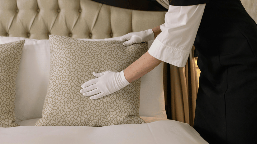 A staff member wearing gloves adjusting a pillow on a neatly made bed with a tufted headboard and curtains visible in the background.