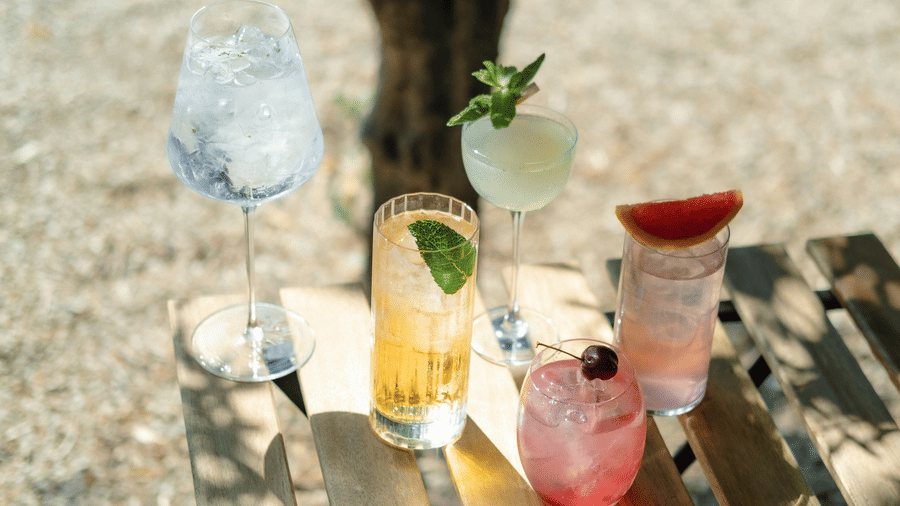 A variety of chilled cocktails in different glassware arranged on a wooden slatted table, garnished with fresh mint, citrus, and a cherry.