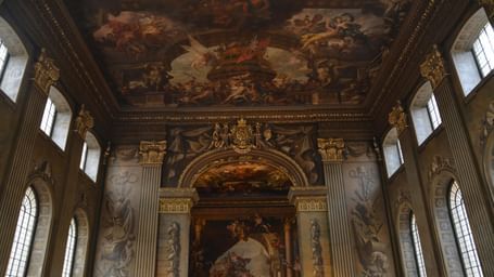An opulent interior view of a grand hall, showcasing a high, vaulted ceiling with a large, classical painting and intricate architectural details on the walls.