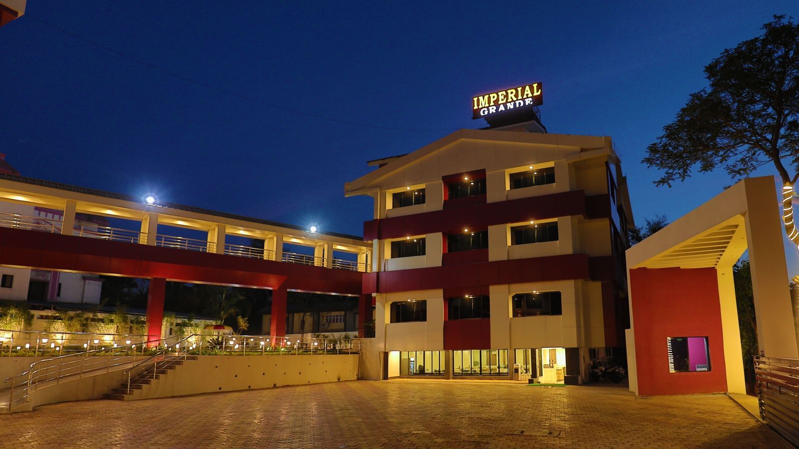 The exterior of Imperial Grande at night, showing a multi-story building with illuminated logo and a paved courtyard reflecting the lights.