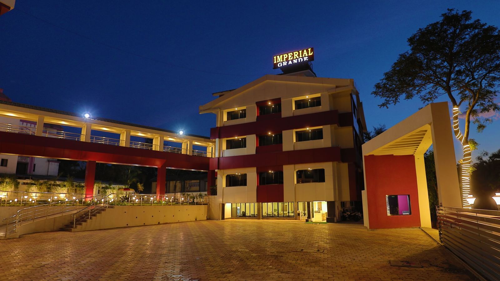 The exterior of Imperial Grande at night, showing a multi-story building with illuminated logo and a paved courtyard reflecting the lights.