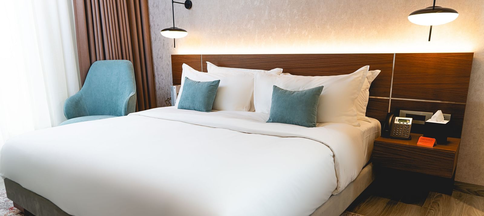 A bed and accent pillows at KertelSuites, featuring a wooden headboard, two bedside tables with lamps, and a armchair by the window