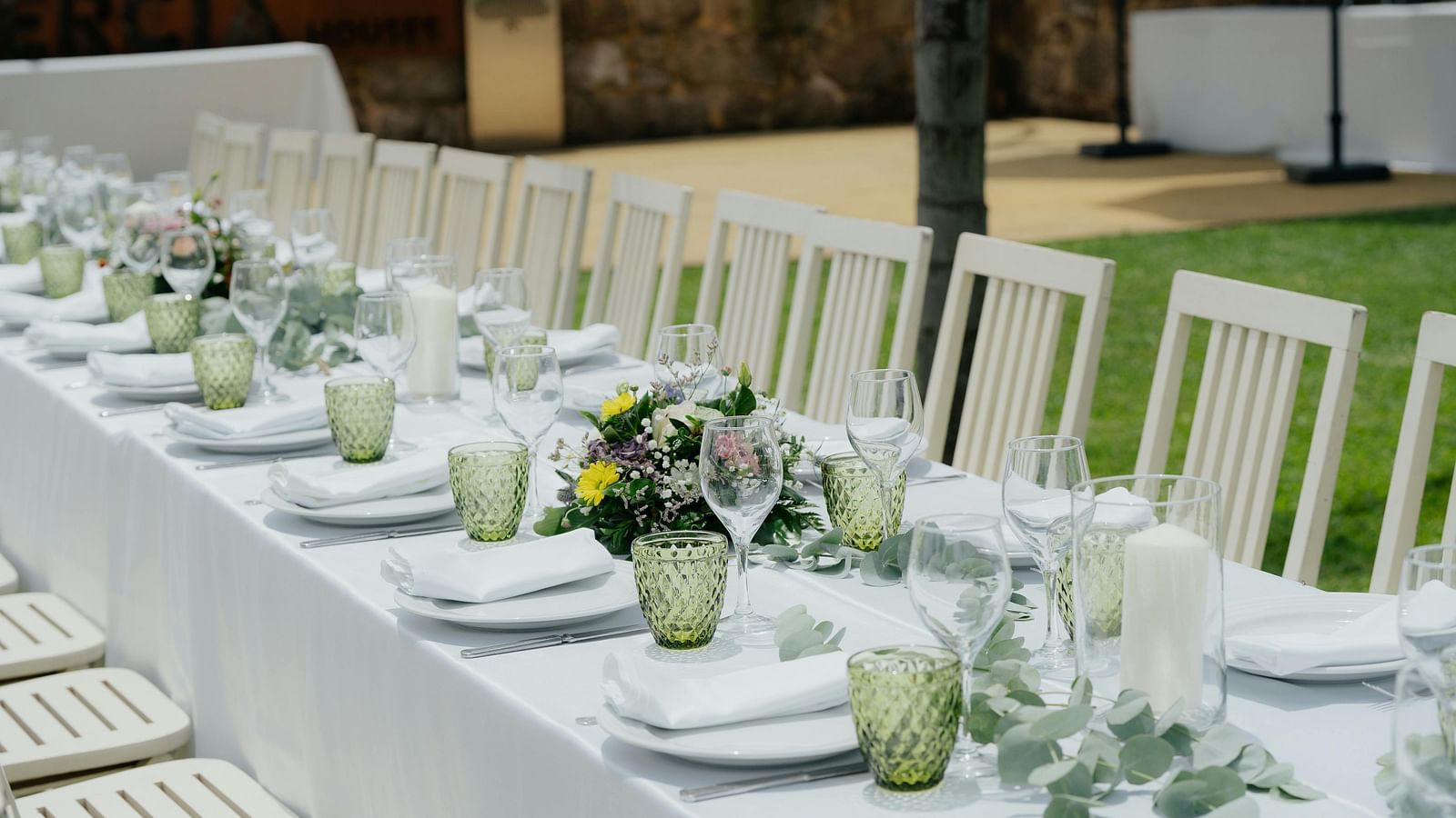 A long outdoor dining table is set with white tablecloths, white chairs, green glasses, and floral centrepieces, ready for a gathering.