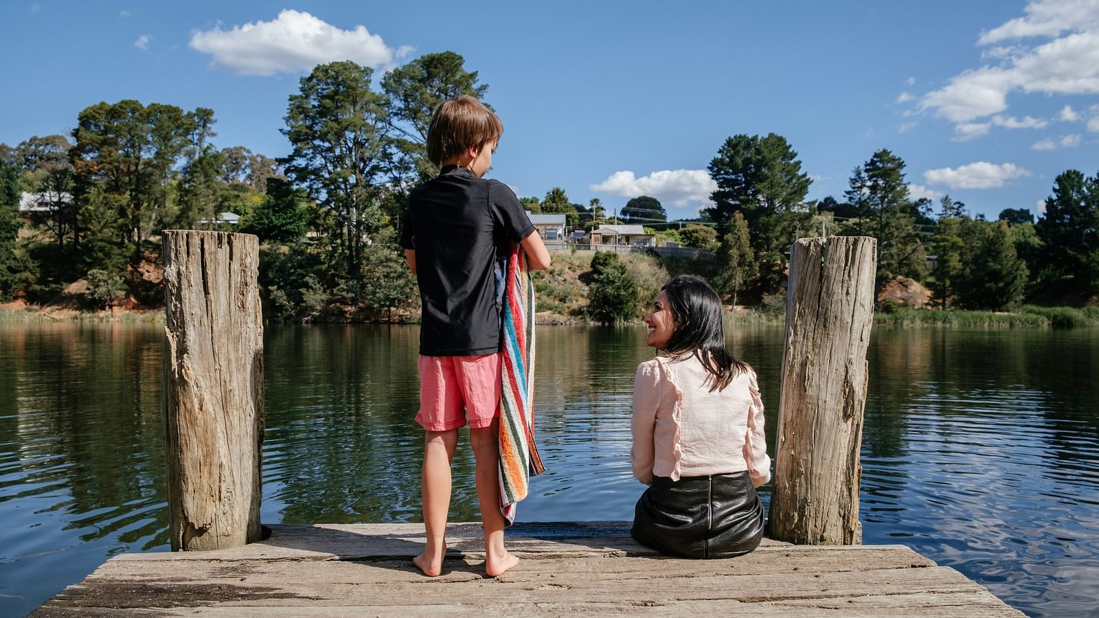 A young boy and girl sitting on a wooden pier overlooking a calm lake surrounded by lush greenery under a bright blue sky.